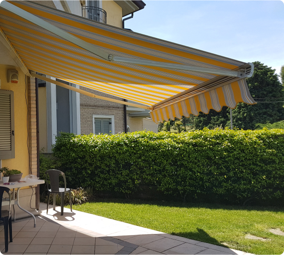 Una tenda da sole gialla e bianca è estesa su un patio, con un giardino verde ben curato sullo sfondo. Una sedia e un tavolo sono visibili.