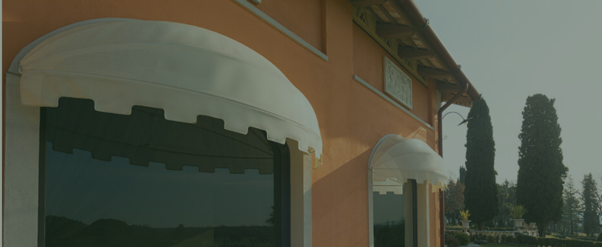 Vista di una facciata di un edificio con intonaco arancione, evidenziata da tende da sole bianche, circondata da vegetazione.