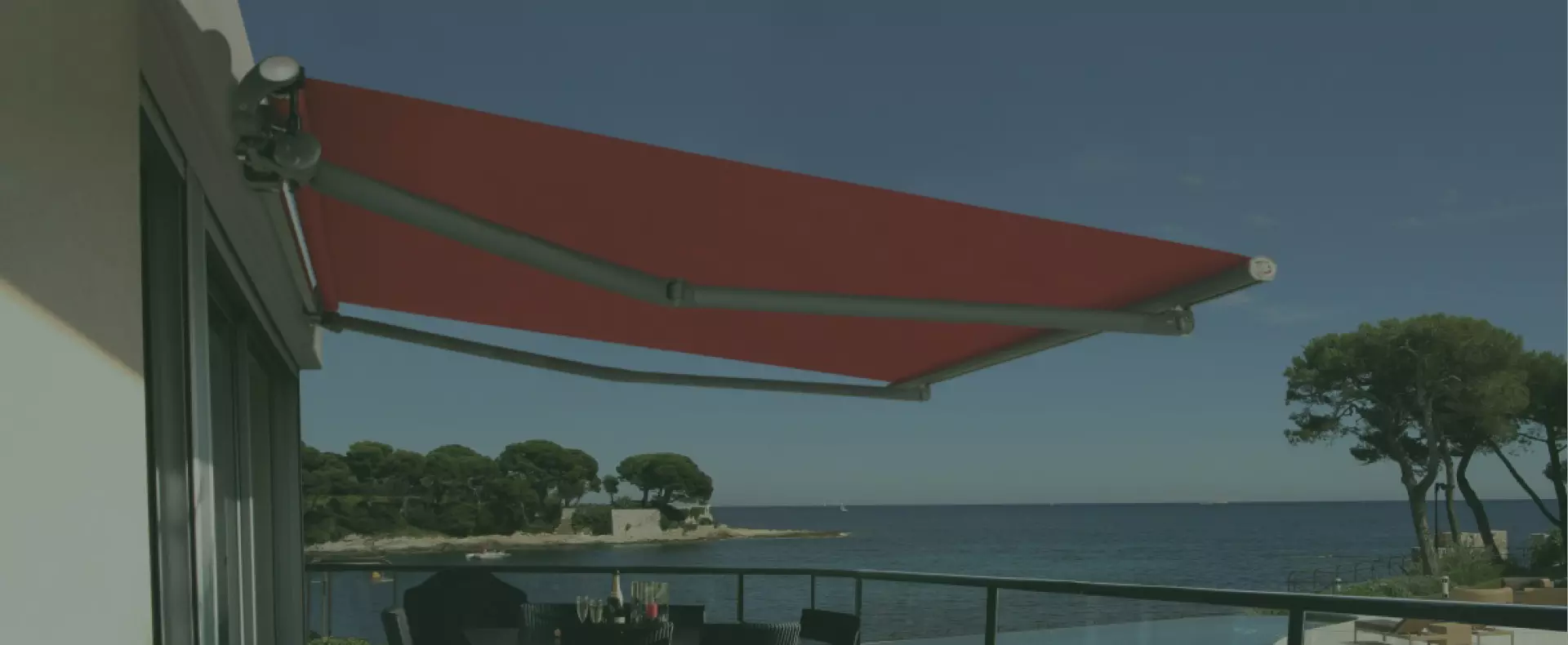 Tenda da sole rossa posizionata sopra un balcone con vista sul mare, su uno sfondo di alberi e cielo sereno.